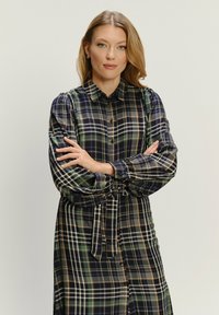 Žena s blond vlasmi, oblečená v dlhých rukávoch gombíkovej kockovanej šaty, so založenými rukami na pozadí s jednoduchým svetlým pozadím.