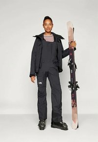 Črna smučarska jakna in kombinezon z teksturiranim burgundskim zgornjim delom, ki drži smučko. Na jakni je logotip North Face, trdi temni smučarski čevlji.