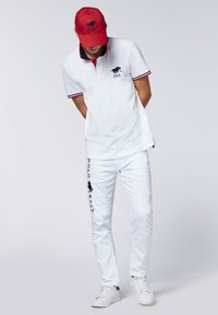 Weißes Polohemd mit blauen und roten Akzenten, gesticktes Logo auf der linken Brust, kombiniert mit weißen Hosen mit seitlichem Logo, abgerundet mit einer roten Mütze.