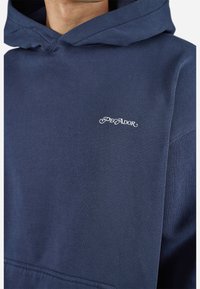 Navy blauwe hoodie met een klein wit "PEGADOR" logo op de borst, gedragen door een persoon met de kap omlaag.