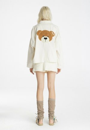 Persona con chaqueta cremosa con un gran parche de osito de peluche en la espalda, pantalones cortos crema, calentadores de piernas color marrón claro y sandalias, de pie de espaldas.