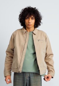 Beige corduroy jacket with a zip front, snap collar, and a chest pocket. Worn over a green crewneck and accompanied by blue denim.