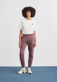 Λευκό μπλουζάκι με μαύρο λογότυπο Nike συνδυασμένο με μπορντό φόρμα. Το μοντέλο φορά ανοιχτόχρωμα αθλητικά παπούτσια και ποζάρει σε μπλε χαλί.