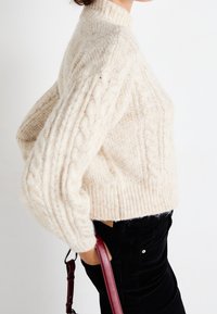 Cremfarbener Grobstrickpullover mit hohem Kragen, gerippten Bündchen und Saum. Getragen mit schwarzen Cordhosen und einer bordeauxroten Umhängetasche.