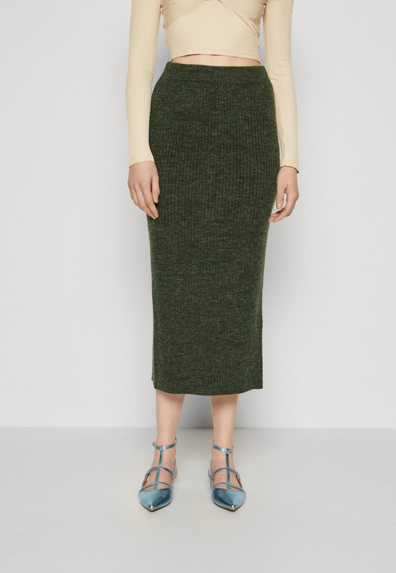 Femme portant une jupe midi en maille côtelée vert foncé taille haute, des talons bleus brillants à bout pointu avec bride à la cheville et un top court beige.