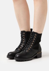 Botas negras hasta el tobillo con una sección de tejido texturizado, ojales dorados, cordones en la parte delantera y tacón cuadrado. Suela de goma con un patrón de relieve resistente.