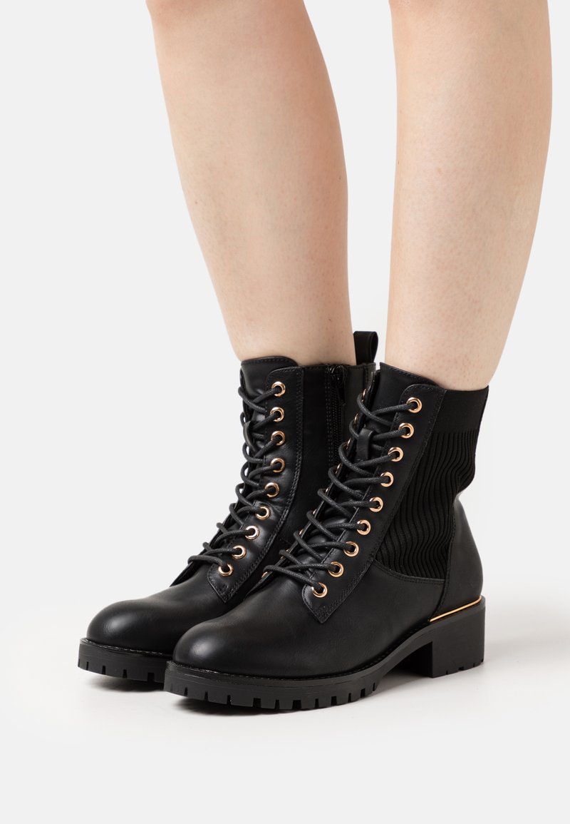 Botas negras hasta el tobillo con una sección de tejido texturizado, ojales dorados, cordones en la parte delantera y tacón cuadrado. Suela de goma con un patrón de relieve resistente.