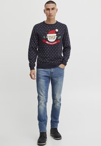 Marineblauer Strickpullover mit weißen Punkten, einem Santa-Gesicht Design und dem Text "Ho! Ho! Ho!"; kombiniert mit ausgebleichten blauen Jeans und schwarzen Stiefeln.