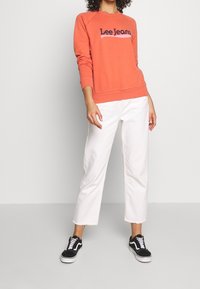 Korálová mikina s logem "Lee Jeans" v kombinaci s bílými širokými džínami. Model nosí černobílé tenisky. Jednoduchý, neformální outfit.