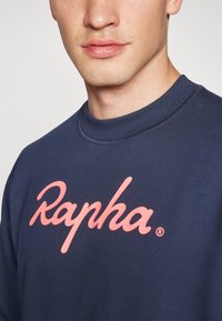 Sudadera navy con cuello redondo, con un gran logo rosa de "Rapha" en el pecho. Textura suave y puños de manga acanalados.