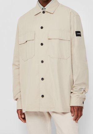 Beiges Langarmhemd mit schwarzen Knöpfen, zwei Brusttaschen mit Klappen und einem schwarzen Calvin Klein Logo-Patch am linken Ärmel.