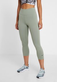 Šedozelené kapri legíny s vysokým pasem, hladkým povrchem a malým černým logem Nike na dolní levé noze. Nosené s atletickými botami.