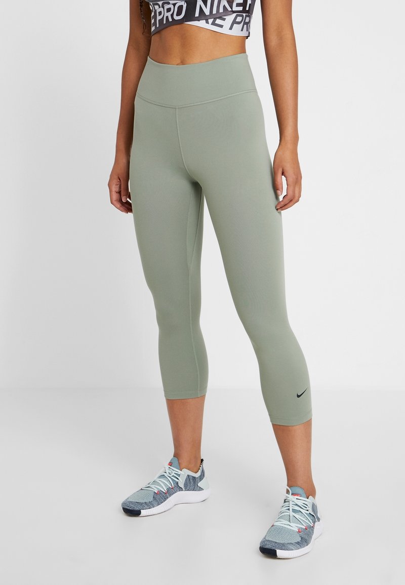 Šedozelené kapri legíny s vysokým pasem, hladkým povrchem a malým černým logem Nike na dolní levé noze. Nosené s atletickými botami.