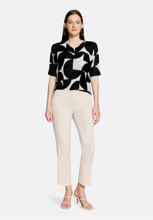 Femme portant un haut à manches courtes à motif abstrait noir et blanc, un pantalon beige clair et des sandales beiges à talons, debout devant un fond uni.