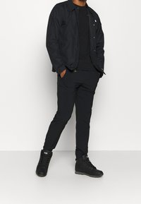 Personne portant une veste noire, un pantalon noir et des chaussures de randonnée Helly Hansen noires, se tenant avec les mains dans les poches sur un fond uni.