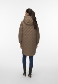 Kvinna med långt mörkt hår i en hästsvans, iklädd en brun vadderad huva, blå tajta jeans och svarta klackade ankelskottar som står framför en vit bakgrund.