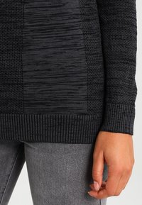 Černý textured pletený svetr s různými vzory a žebrovaným lemem. Ruka modelky viditelná, spočívající na šedých džínách.