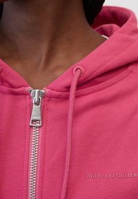 Primer plano del cuello y pecho de una persona que lleva puesta una chaqueta con cremallera rosa y tirantes plateados, mostrando el texto "Marc O’Polo DENIM".