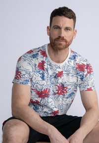 Camiseta de manga corta, ajustada, hecha de algodón con una base blanca y patrones florales en rojo y azul, con cuello redondo.