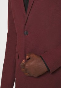 Bordowa marynarka z pojedynczym guzikiem, bocznymi kieszeniami, teksturowaną tkaniną i czarną podszewką na mankietach, przedstawiona z ręką poprawiającą klapę.