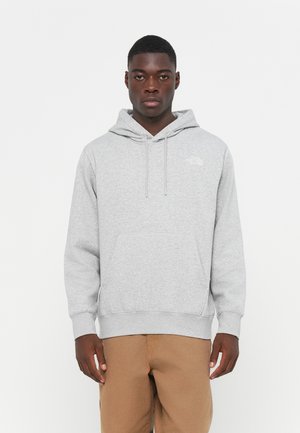 Sudadera gris de tela suave, con un bolsillo frontal, capucha con cordón y un pequeño logo blanco en el pecho. Combinada con pantalones color beige.