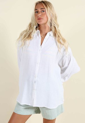 Femme blonde aux cheveux ondulés portant une chemise blanche ample à boutons et un short vert clair sur un fond beige.