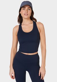 Vrouw in een marineblauwe sportcrop top, hoge tailleleggings en een grijze honkbalpet, staand tegen een effen lichte achtergrond.