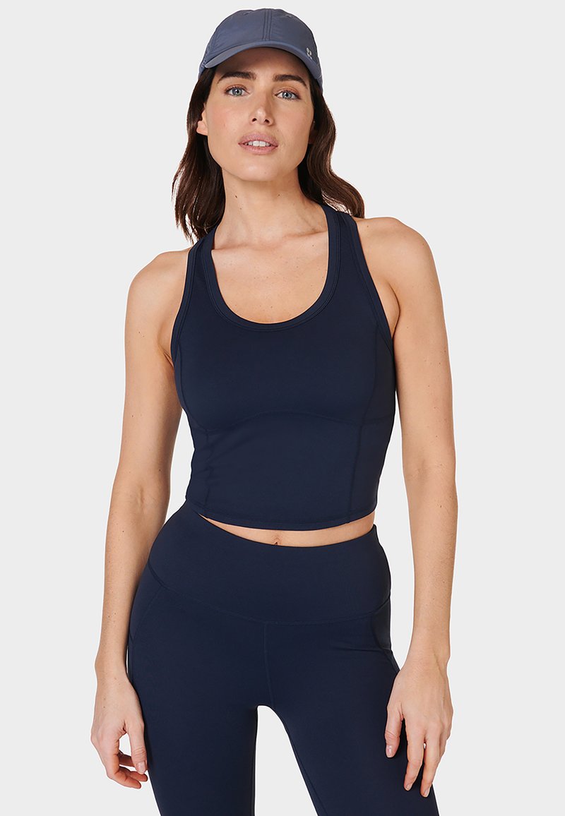 Vrouw in een marineblauwe sportcrop top, hoge tailleleggings en een grijze honkbalpet, staand tegen een effen lichte achtergrond.