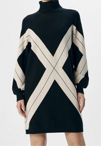 Zwart en crèmekleurige gebreide jurk met een hoge col, voorzien van geometrische patronen en losse mouwen, die boven de knie eindigt.