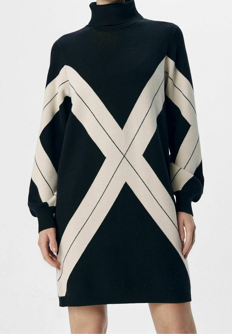 Zwart en crèmekleurige gebreide jurk met een hoge col, voorzien van geometrische patronen en losse mouwen, die boven de knie eindigt.