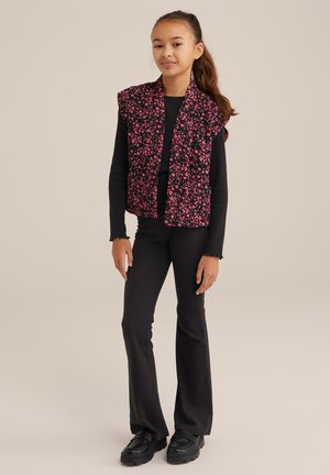 Blumengemusterte gesteppte Weste in Schwarz mit pinken Blumen, kombiniert mit einem langärmeligen schwarzen Oberteil und schwarzen Schlaghosen. Schwarze Schuhe mit Gummisohle.