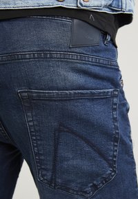 Los jeans de mezclilla de un azul oscuro presentan un bolsillo trasero, una tela texturizada y un parche de marca de cuero. El detalle de la costura añade un interés visual.