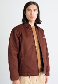 Brun, zip-up jacka med krage, tillverkad av texturerat tyg; har sidofickor och subtil logotypdetalj på bröstet.