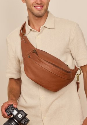 Lächelnder Mann, der ein beiges Hemd und eine braune Leder-Umhängetasche trägt, hält eine schwarze DSLR-Kamera in der rechten Hand vor einem einfarbigen Hintergrund.