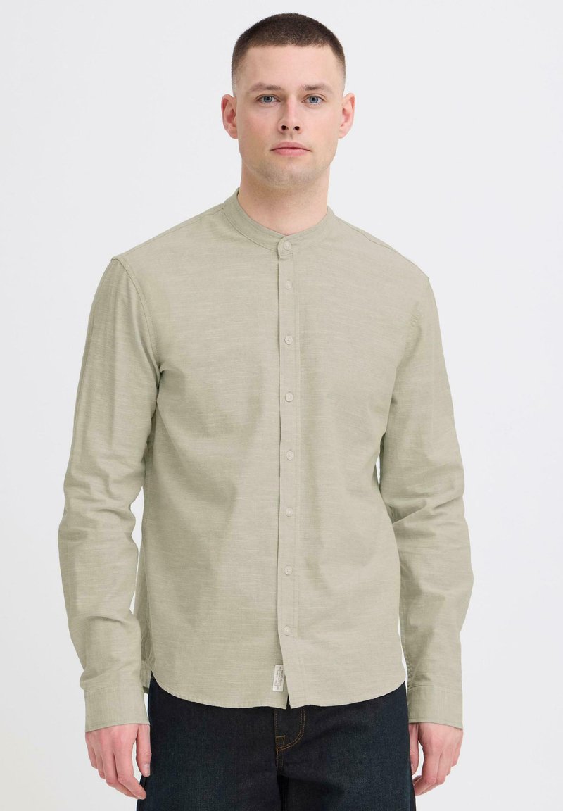 Jeune homme aux cheveux courts portant une chemise beige clair à col mandarin boutonnée et un pantalon foncé, debout devant un fond blanc.