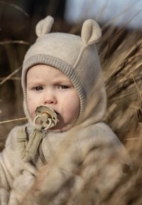 Beiger Fleece-Hoodie mit Bärenohren, ausgestattet mit einem Reißverschluss und einem Clip für einen Schnuller. Weiche Textur, getragen über einem hellfarbenen Kleidungsstück.