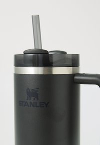 Tazza in acciaio inossidabile nera con coperchio trasparente, cannuccia in plastica grigia e manico laterale. Presenta il logo Stanley in blu scuro.