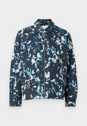 Jaqueta de camuflagem azul e preta, fabricada em tecido leve, com fecho na frente, colarinho tipo ponto e um patch de logotipo na parte inferior da frente. Punhos e barra elásticos.