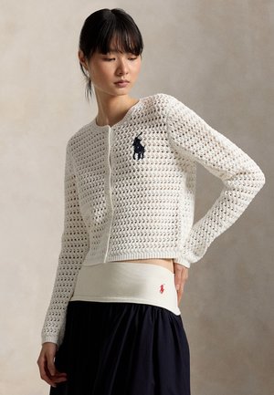 Kobieta ubrana w biały dzianinowy kardigan z granatowym logo oraz czarną spódnicę z białym paskiem w talii ozdobionym małym czerwonym logo.