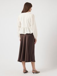 Kvinne med brunt hår i en hvit langermet peplum-bluse, brun midi-skjørt og grå kitten heel slingback-sko, som står med ryggen til.