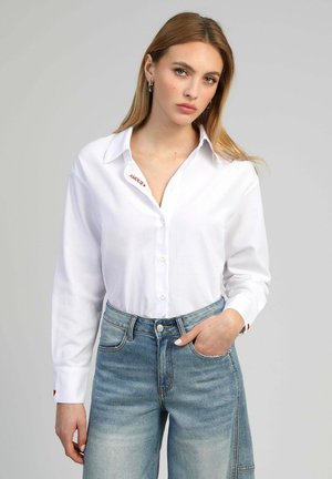 Jeune femme aux longs cheveux blonds portant une chemise blanche boutonnée et un jean taille haute bleu clair, debout avec une main dans la poche.