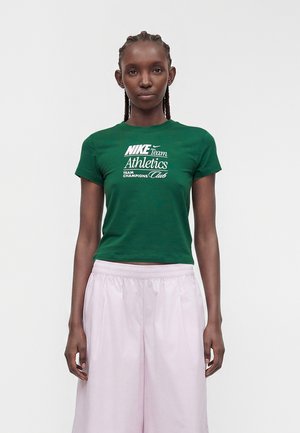 Zöld Nike Team Athletics pólót és világos rózsaszín bő szárú nadrágot viselő, fonott hajú nő áll egyszínű háttér előtt.
