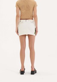 Gonna mini in denim bianco con orlo grezzo, due bottoni circolari in vita e dettagli ricamati verdi sul lato. Design svasato.