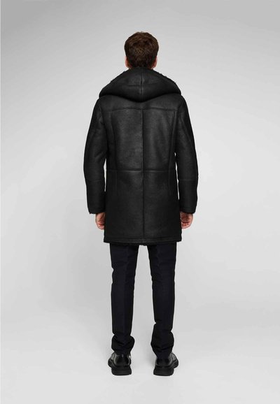 Uomo in piedi con la schiena rivolta, indossando un cappotto in shearling nero con cappuccio, pantaloni neri e scarpe nere su uno sfondo semplice.