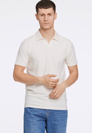 Polo shirt beige clair en coton, avec un col classique et des manches courtes, assorti à un jean en denim bleu. Design simple et épuré.