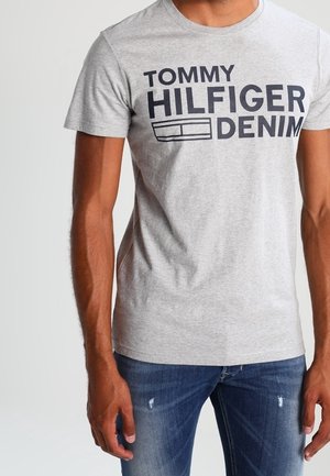 Mann trägt ein hellgraues Tommy Hilfiger Denim T-Shirt und ausgewaschene blaue Jeans, steht vor einem schlichten Hintergrund.