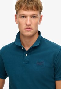 Polo in teal realizzato in cotone testurizzato, con bavero pieghettato, chiusura con tre bottoni e un piccolo logo ricamato sul petto.