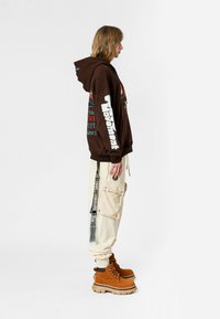 Persona de perfil lateral vistiendo una sudadera gráfica marrón, pantalones cargo crema con parches y botas de plataforma en color beige con cordones, sobre fondo blanco.