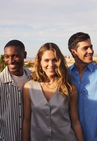 Trois personnes sourient, vêtues de tenues décontractées : une veste à boutons rayée, un haut sans manches gris clair et une chemise à col bleue.