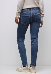 Vaqueros ajustados de mezclilla azul con bolsillos traseros, ligero desgaste y cintura media, combinados con zapatillas blancas y un suéter claro.